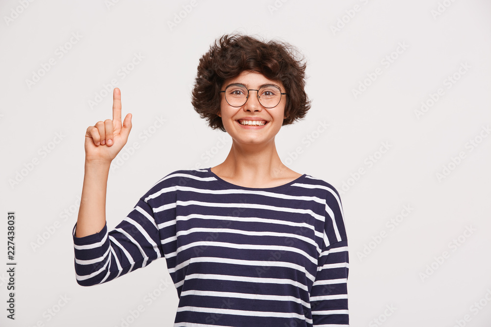 Portrait of smiling happy young woman with curly brown hair, wears white and blue stripped sweatshirt and transparent black-framed round glasses points up with finger isolated over white background Portrait of smiling happy young woman with curly brown hair, wears white and blue stripped sweatshirt and transparent black-framed round glasses points up with finger isolated over white background