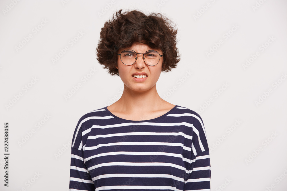 Portrait of young woman with curly brown hair, wears white and blue stripped sweatshirt and transparent black-framed round glasses feels disgust wrinkles nose isolated over white background Portrait of young woman with curly brown hair, wears white and blue stripped sweatshirt and transparent black-framed round glasses feels disgust wrinkles nose isolated over white background
