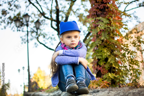 little girl in a blue hat sits and sad autumn sunny day