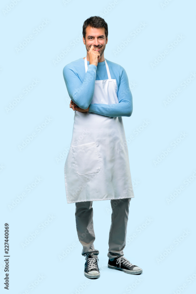 Full body of Man wearing an apron smiling with a sweet expression on blue background Full body of Man wearing an apron smiling with a sweet expression on blue background