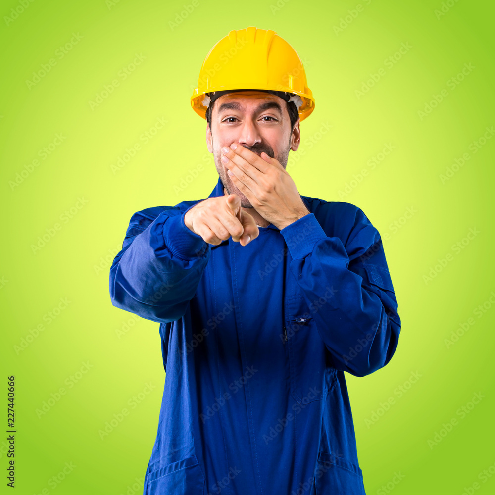Young workman with helmet pointing with finger at someone and laughing a lot while covering mouth on green background