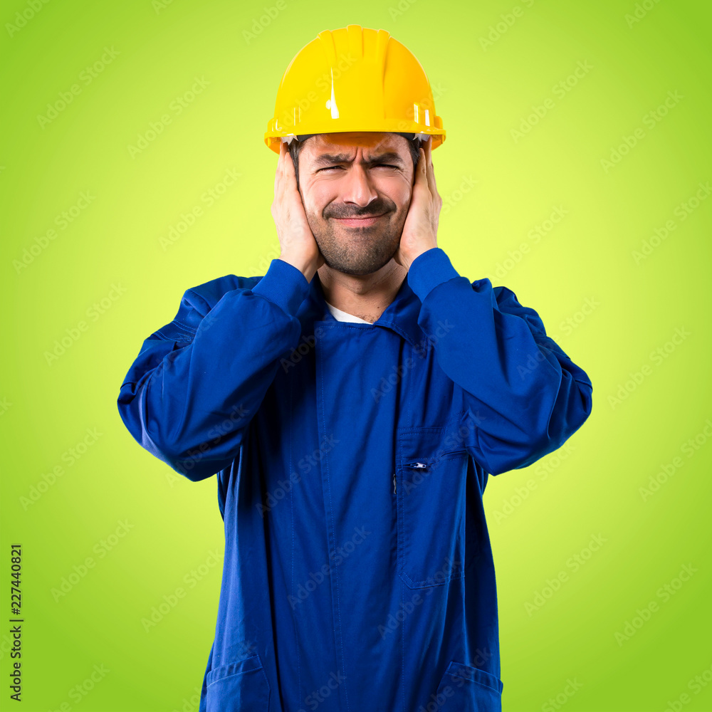 Young workman with helmet covering both ears with hands. Frustrated expression on green background
