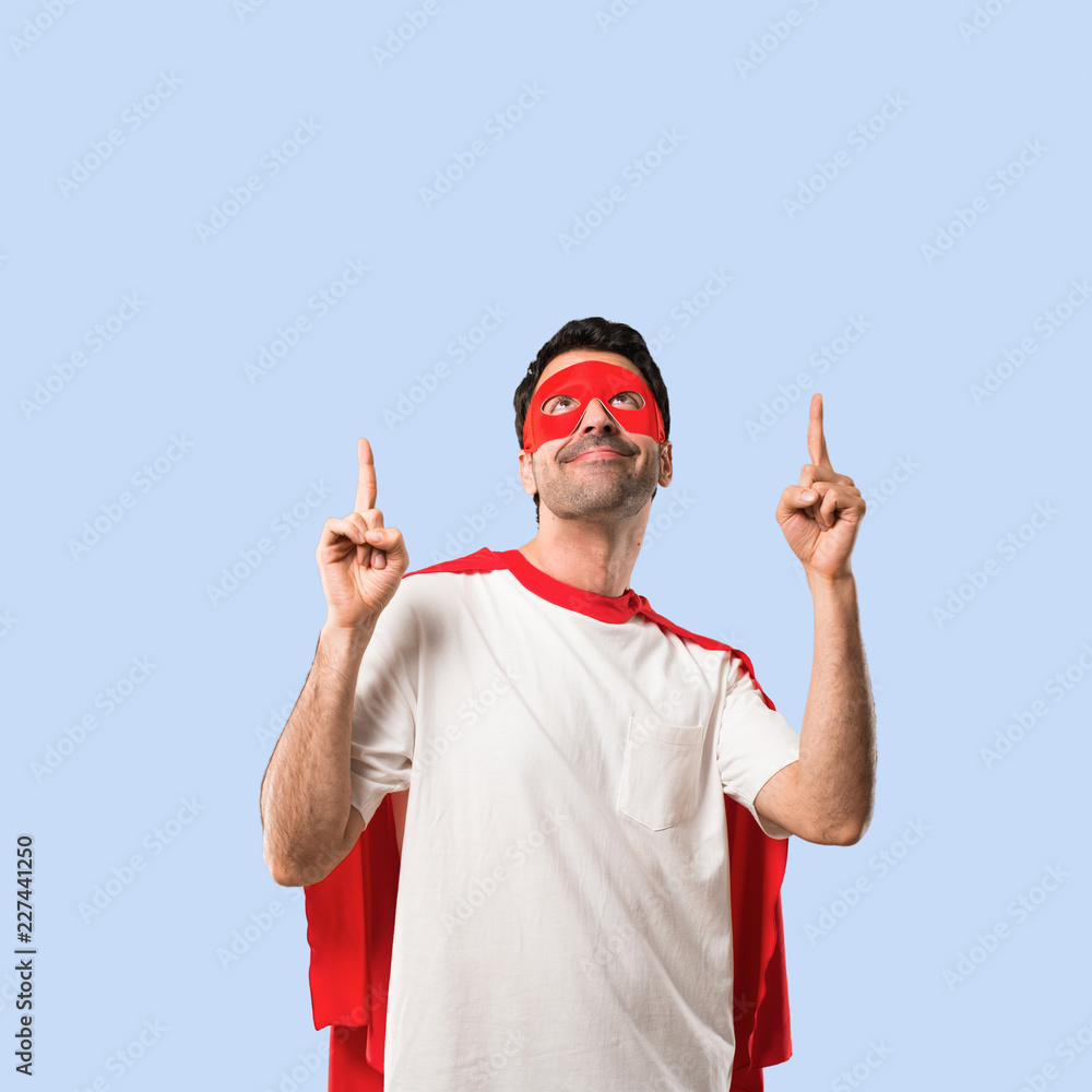 Fototapeta premium Superhero man with mask and red cape pointing with the index finger a great idea and looking up on isolated blue background