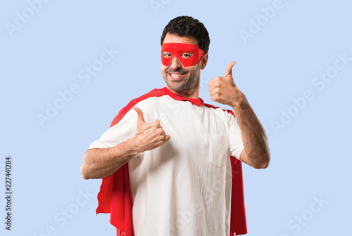 Superhero man with mask and red cape giving a thumbs up gesture and smiling because has had success on isolated blue background