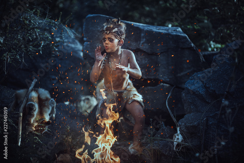 Fototapeta Naklejka Na Ścianę i Meble -  Shaman boy at the fire. Scary young primitive boy outdoors near bonfire. Witch craft concept. Angry caveman, manly boy with horns near bonfire. Prehistoric tribal man outdoors on nature making
