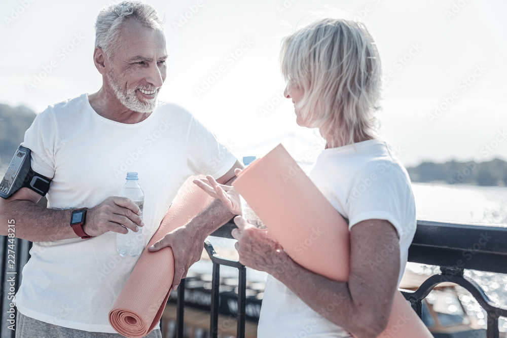 Fototapeta premium Healthy people. Pleased bearded male person holding bottle in right hand and having case for telephone on arm, looking at his wife
