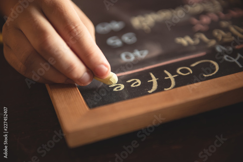 The girl is writing a coffee menu on the board with her right hand using a yellow chalk.