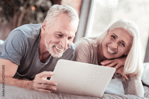 Pleasant emotions. Close up of elderly family expressing delight while lying on the bed and using a laptop