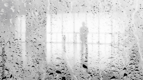 Blurred silhouette of man in the light hall through large wet glass wall. Black and white image
