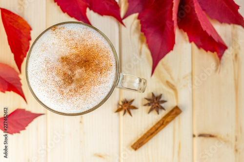 Hot drink, coffee latte or cappucino with cinnamon on milk foam in a glass mug, spices and red autumn leaves on wooden background, cold season concept, top wiew