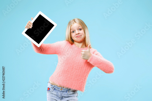 Little funny girl with tablet on blue studio background. She showing something and pointing at screen.