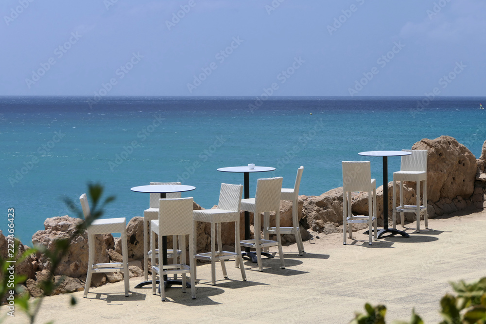 Relaxation - Some bistro tables stand on a path with white stools. In the background you can see the sea with different shades of blue. The sky is cloudless and it is best summer weather. 