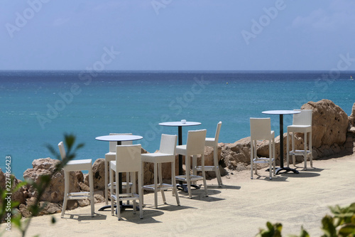 Relaxation - Some bistro tables stand on a path with white stools. In the background you can see the sea with different shades of blue. The sky is cloudless and it is best summer weather.  © Fahrradauszeit