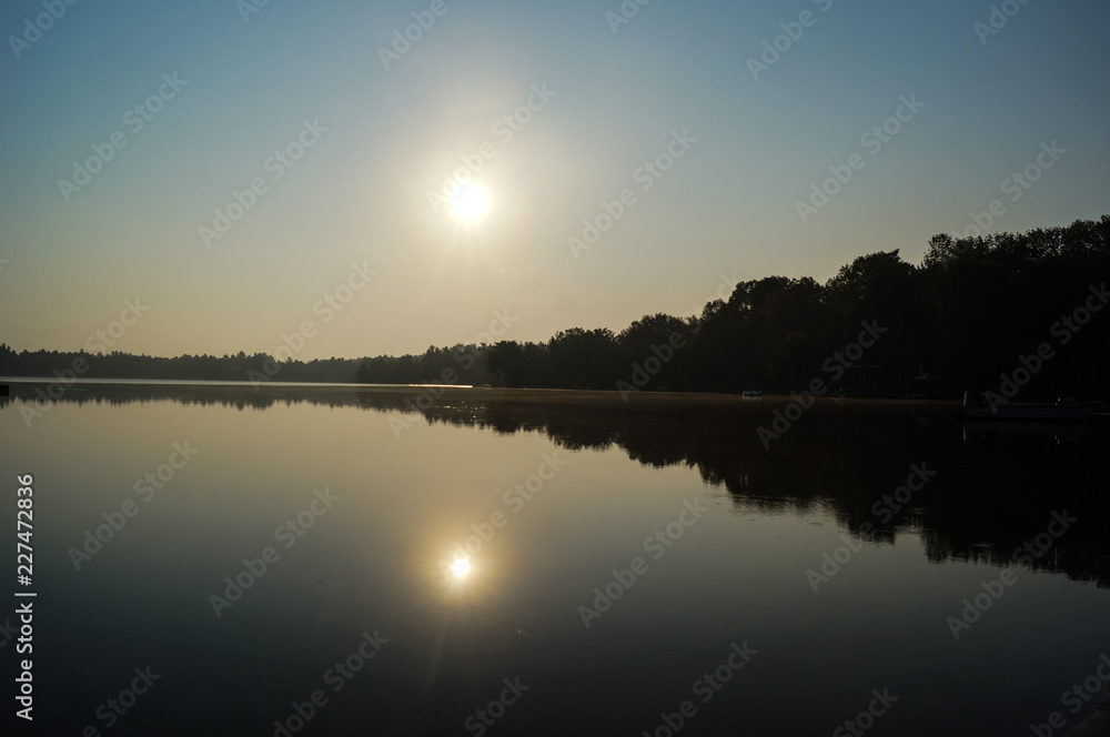 Fototapeta premium Sonnenaufgang spiegelt sich im See vor anwachsender Baumkette