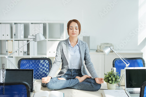 Young businesswoman with eyes closed sitting on her office table and meditating during break