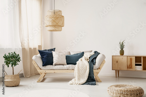 Light lounge with pillows and two blankets placed in white sitting room interior with window with curtains, straw lamp and fresh plant on floor in real photo