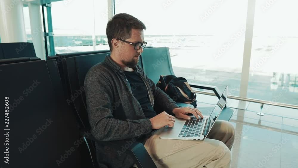 Adult bearded male passenger is working with notebook, sitting in a waiting hall of airport. He is pressing buttons on keyboard and using touchpad Adult bearded male passenger is working with notebook, sitting in a waiting hall of airport. He is pressing buttons on keyboard and using touchpad