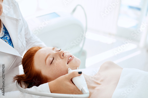 Apparatus face treatment in medical spa center, beautiful redhead lady receiving electric facial peeling massage. Cosmetological methods, apparatus cosmetology, SPA-procedures Concept. photo