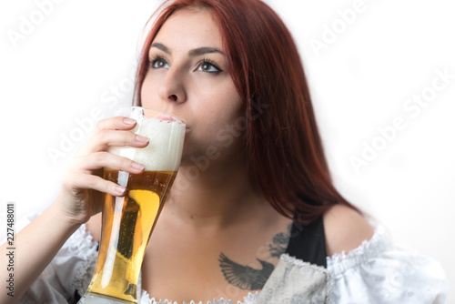 A young girl with long red hair in a traditional German corset drinks a golden beer from a glass
