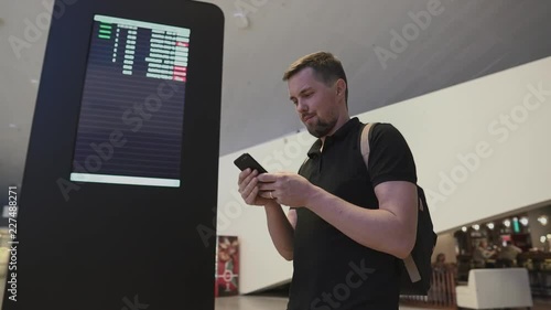 Portrait of a handsome man with backpack using smartphone by digital information board. Smiling man sms texting indoor.