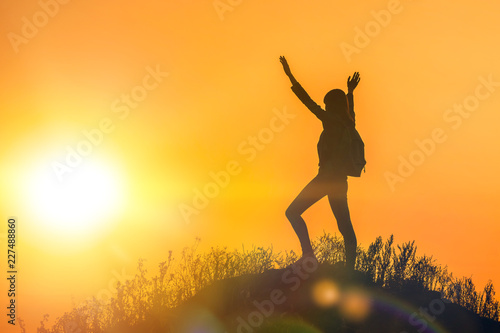 silhouette of a young happy girl with a backpack holding her hands up the sportswoman  on top of the mountain in the glare of the sun in the sunset. Fitness  victory