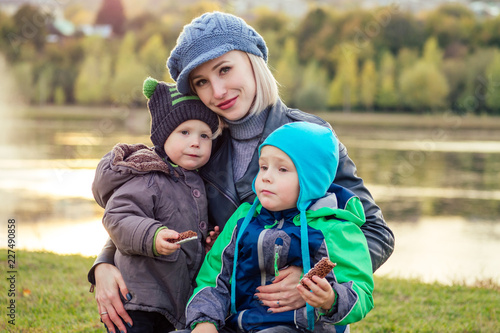 happy and beautiful blonde woman in hat with two baby son eating a chocolate bar in a warm jacket in the autumn park against the backdrop of the lake. stylish mother with child tasty snack picnic © yurakrasil
