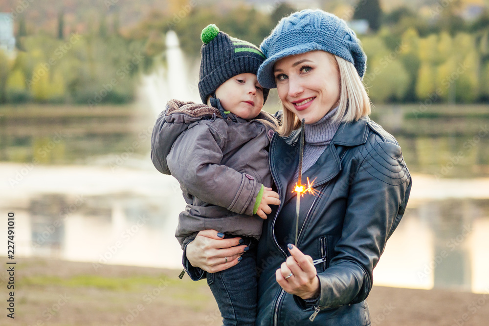 Mother in a warm jacket and knitted hat hugging her baby boy walking in the autumn park. stylish blondewoman with a child against the backdrop of the lake Mother in a warm jacket and knitted hat hugging her baby boy walking in the autumn park. stylish blondewoman with a child against the backdrop of the lake