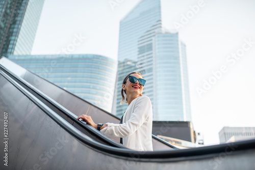 Stylish businesswoman in white suit going up on the escalator at the business centre outdoors with skyscrapers on the background in Paris