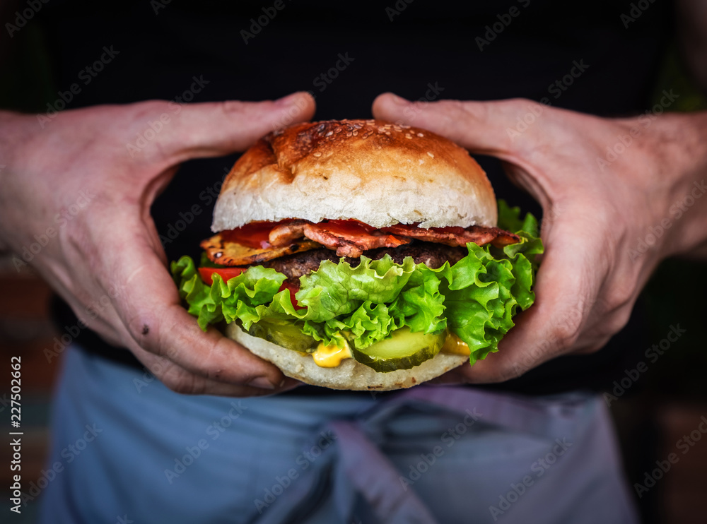 Male hands holding a juicy tasty cheeseburger with beef, lettuce, pickles, tomato and onion rings on a wooden table. Classic street food - grilled burger