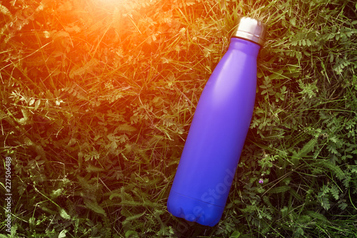 Stainless thermos water bottle, matte blue color. Mockup isolated on green grass background with sunlight effect.
