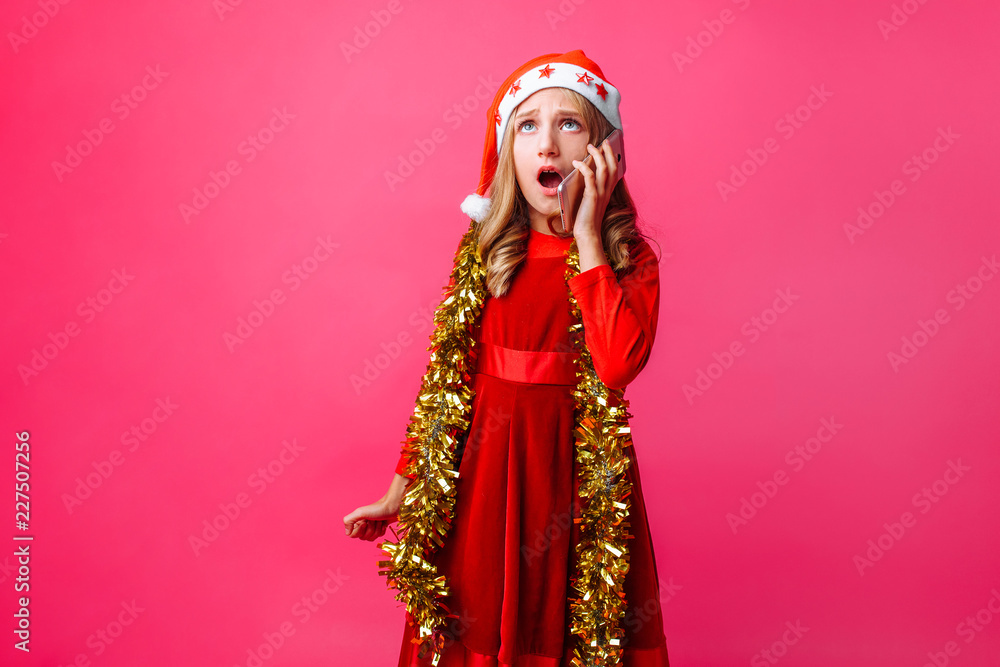 Portrait of schoolgirl girl in Santa hat and tinsel on neck, emotionally talking on phone with surprised facial expression isolated on red background