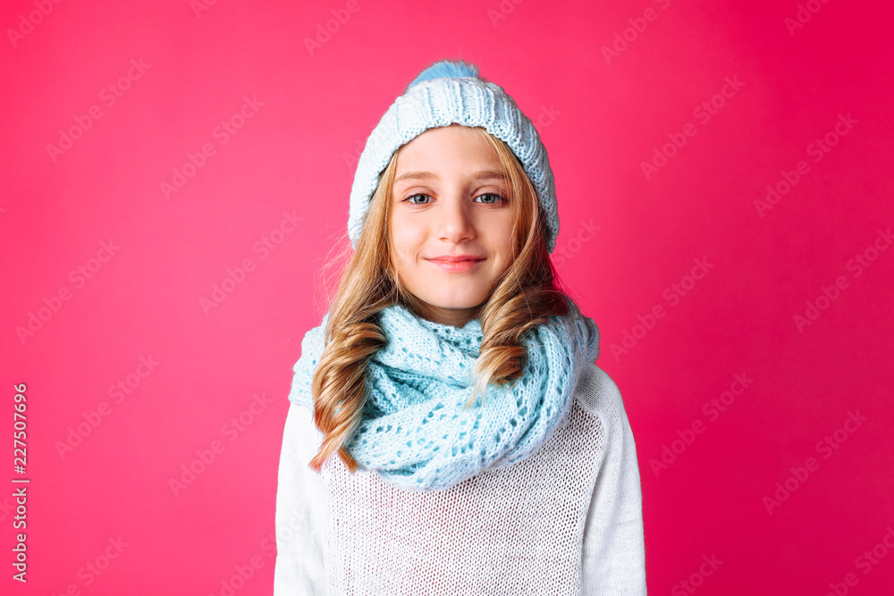 cute teenage girl in white sweater standing isolated on pink background wearing warm blue hat and warm scarf.