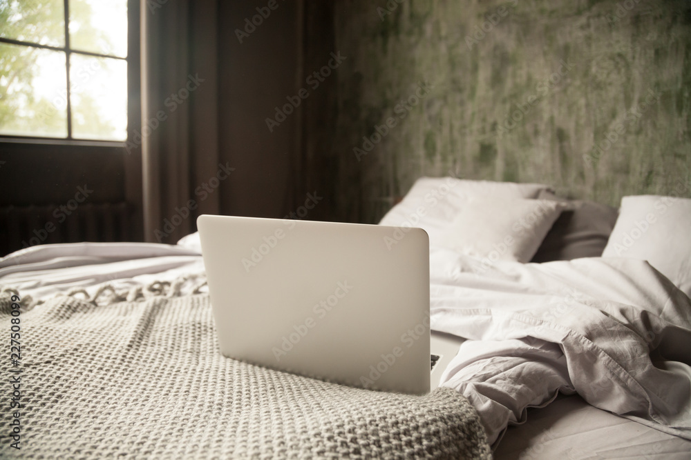 Fototapeta premium Empty cozy room with an open computer on a bed in bedroom at home no people. Morning light through the window. Weekend and working at home, leisure and lifestyle, wireless technology concept, close up
