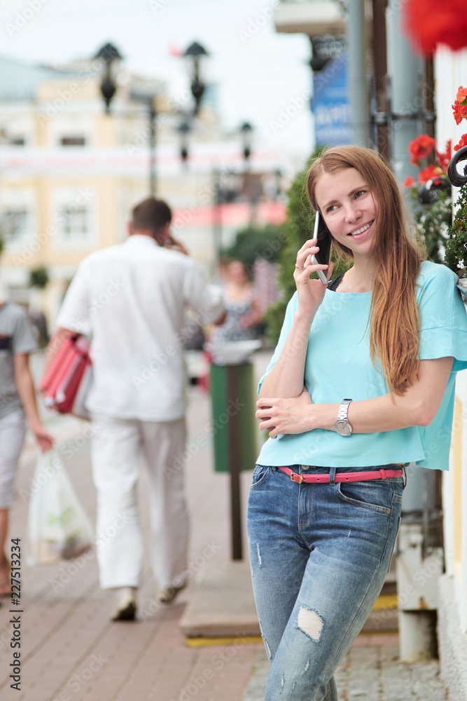Fototapeta premium Young skinny redhead woman stands in the street, smiles and has a conversation by phone
