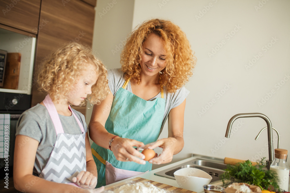 Little cooker at work. Fair haired six years old girl and her mother kneads the dough from flour standing at the kitchen table. Cooking together, Family concept Little cooker at work. Fair haired six years old girl and her mother kneads the dough from flour standing at the kitchen table. Cooking together, Family concept