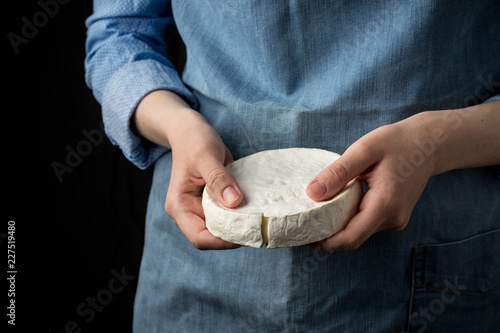 Woman in blue apron holding soft french camembert cheese on dark background