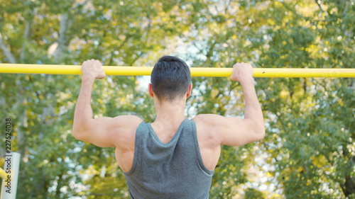 Young muscular man doing pull up exercise on horizontal bar. Slim athlete a very fit guy fitness instructor or a personal trainer working out his arm muscles on outdoor gym as part crossfit workout.