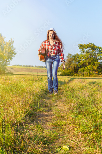 Beautiful, happy, red-haired girl in a field on a sunset background with a guitar in the summer. The concept of relaxation and creative travel