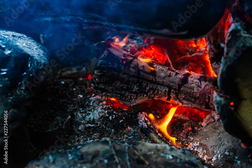 Smoldered logs burned in vivid fire. Atmospheric background with orange flame of campfire. Unimaginable detailed image of bonfire from inside with copy space. Smoke and ashes close up.