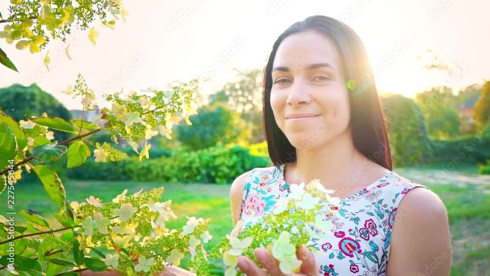 Portrait of a smiling brunette with dimpled cheeks against a green park. Beautiful young woman stands in park and looks at camera. Pretty girl in summer dress with floral print inhales the scent of a Portrait of a smiling brunette with dimpled cheeks against a green park. Beautiful young woman stands in park and looks at camera. Pretty girl in summer dress with floral print inhales the scent of a