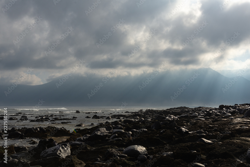the sun's rays against the backdrop of the mountains and the lava-rocky wet shore in the foreground the sun's rays against the backdrop of the mountains and the lava-rocky wet shore in the foreground