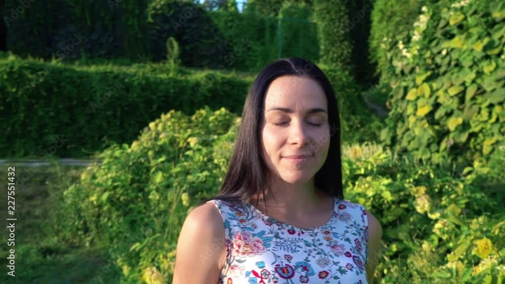 Portrait of a beautiful young woman with dimpled cheeks against a green park. Smiling brunette slowly walks along green bushes in the garden. Pretty girl in summer dress with floral print Portrait of a beautiful young woman with dimpled cheeks against a green park. Smiling brunette slowly walks along green bushes in the garden. Pretty girl in summer dress with floral print