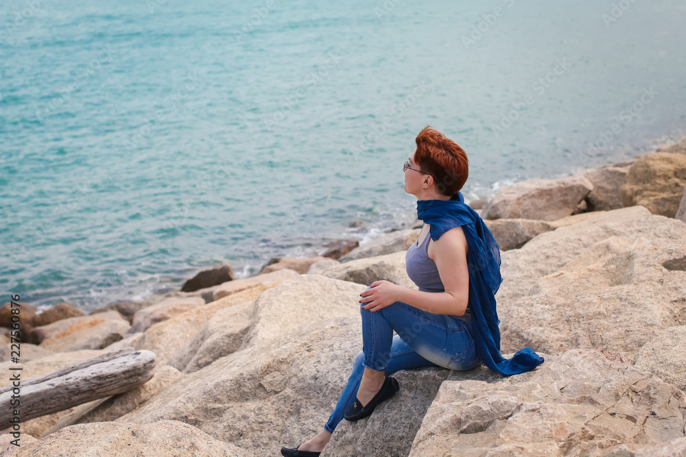 Adult caucasian women sit on rocky beach with blue neckchief relaxing and thinking about something