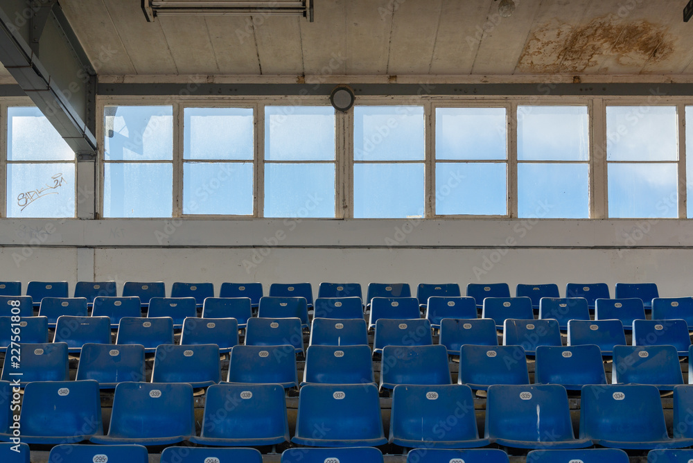 Naklejka premium Front view row of blue plastic seats on rough concrete step floor in old vintage dirty concrete structural stadium with windows frame background without people. 