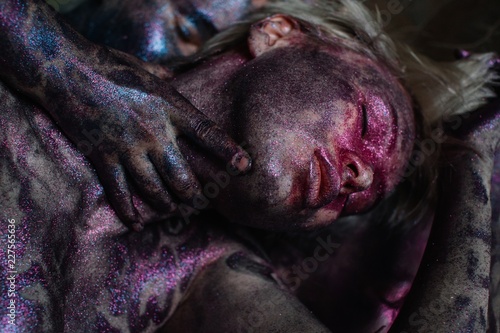 Young woman with violet body art and dreamy closed eyes laying on reflecting background and crop hands touching her neck photo