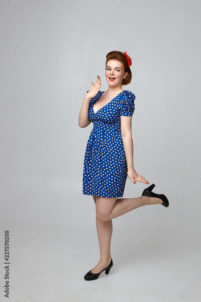 Wow. Vertical picture of attractive fashionable female model wearing 60s  retro outfit having excited facial expression, covering mouth and lifting  up one leg. Flirty pin up girl posing in studio foto de