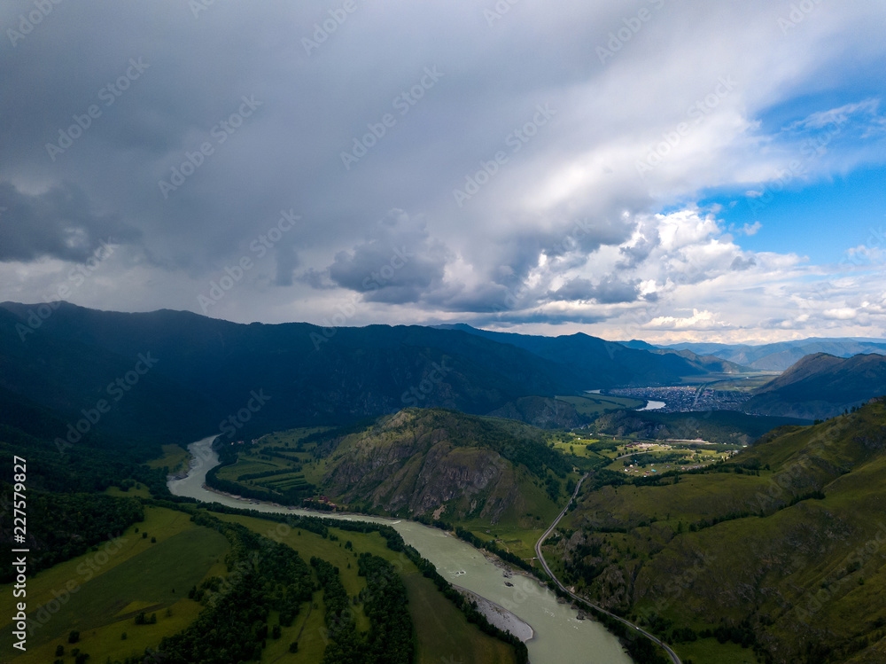 Aerial view of the nature of the Altai Mountains during an approaching thunderstorm with clouds and, as yet, a bright shining on the village at the foot of the mountain with a green meandering river