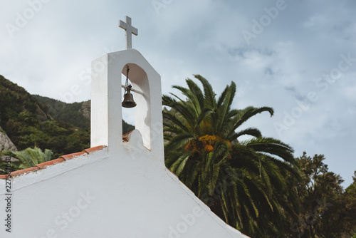 A roof of a small white chapel with its bell and cross and palm trees in a background photo