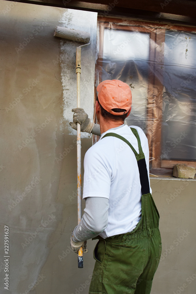 Worker in overalls and cap painting primer on wall with roller before plastering. Facade thermal insulation and painting works during exterior renovation. Home improvement and DIY concept. Worker in overalls and cap painting primer on wall with roller before plastering. Facade thermal insulation and painting works during exterior renovation. Home improvement and DIY concept.