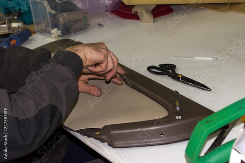 A male worker does manual work on skinning a genuine leather of a vehicle's interior with a beige-colored door to carry out a design solution in a car repair shop at the workplace with scissors